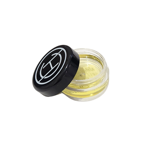 A small open jar of natural oil primer with a black lid, showing yellowish liquid inside. The product is intended for makeup use and the image captures the texture and color of the primer.