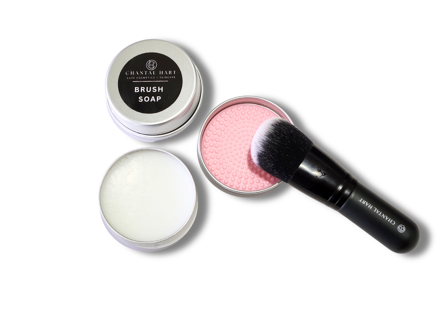 A makeup brush cleaner kit which includes a metal tin of brush soap, a pink lid for the tin, and a black makeup brush being cleaned with the product.
