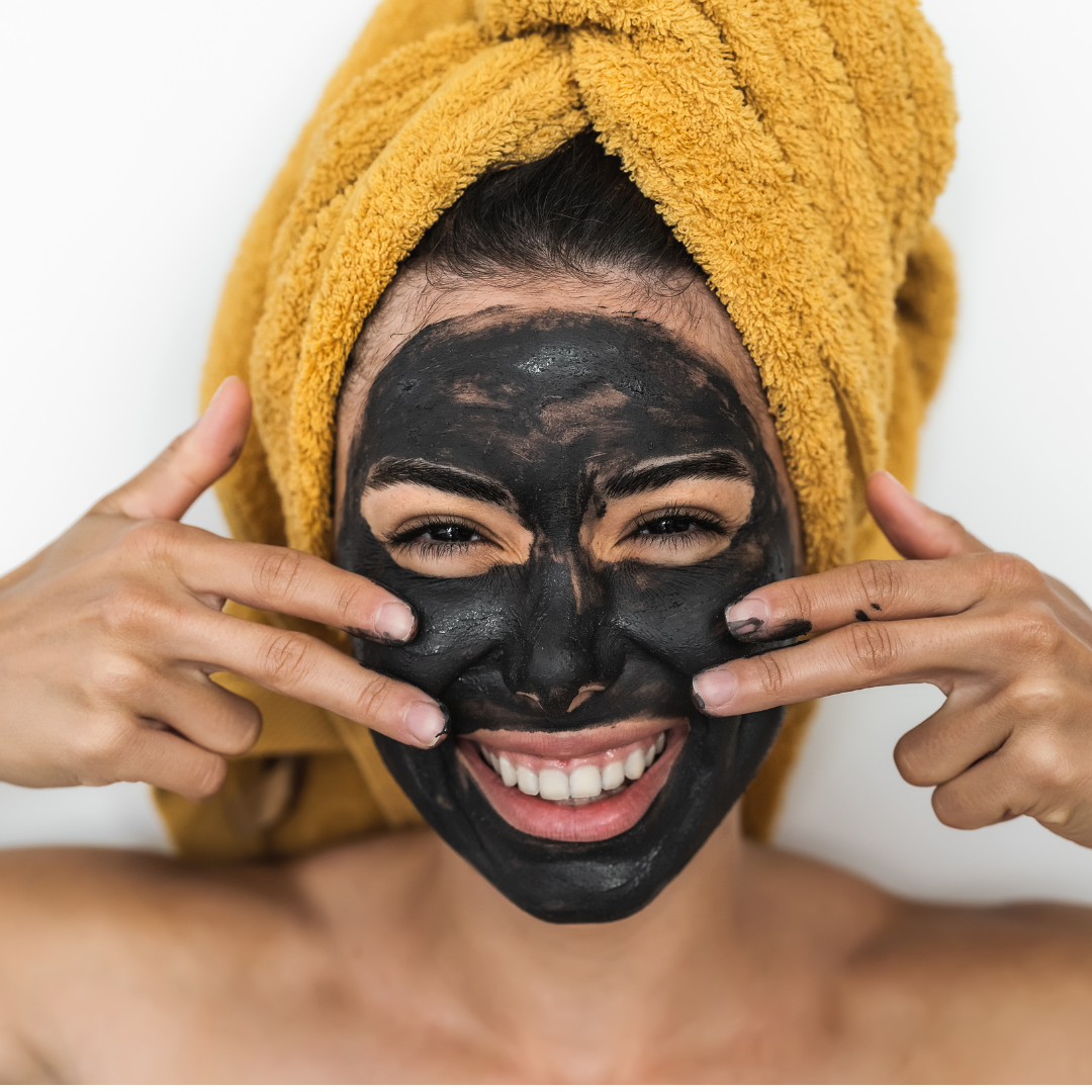 Woman with a charcoal face mask and yellow towel on her head, smiling.