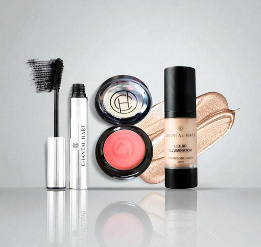 Makeup products including a blush, mascara, and liquid illuminator on a reflective surface with a gray background.